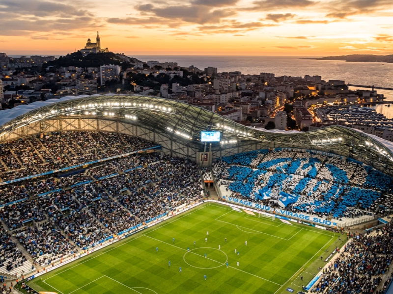Vue aérienne du Stade Vélodrome Marseille avec drapeaux ciel et blanc formant une chorégraphie tribune et Notre-Dame de la Garde à l'horizon
